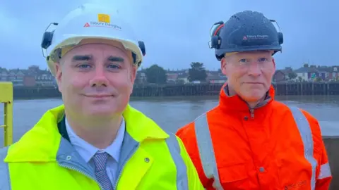 Andrew Turner/BBC Daniel Candon, left in yellow high visibility jacket and Steve Kirk, right, in orange high visibility jacket. Both are wearing hard hats, standing near the River Yare