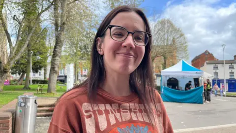 A woman standing in Redditch town centre, wearing a light brown jumper - she is smiling, and in the background are some market stalls.