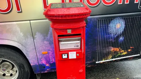 The photo shows a bright red postbox at the centre. It looks like a traditional postbox, except from it has a drawer where small parcels can be posted. There is also a solar panel covering the top of it. It is situated at the side of the road, and a bus is parked behind it.