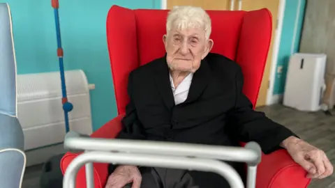 Old man with short grey hair in black blazer and white shirt sat in red chair 