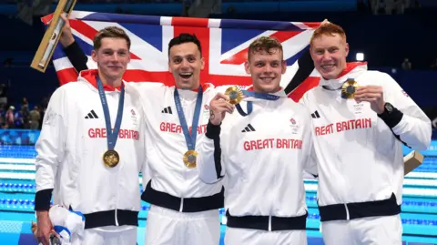 PA Media Duncan Scott, James Guy, Matthew Richards and Tom Dean. Duncan has short light hair, James has short dark hair, Matthew has short light hair and Tom has red short hair. They are all wearing white Adidas branded Great Britain jackets and wearing gold medals. They are holding a union jack behind them and smiling at the camera. 