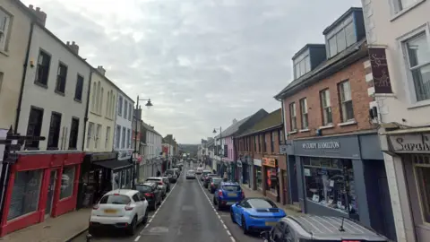 Google Maps A street view image of Ballymoney town, there are numerous cars parked on either side of the street. There are a range of different shops painted in grey, orange, red and black.