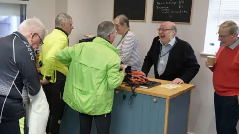 Three men wearing cycling clothes approach a shop counter, where a man and a woman are serving with smiles on their faces. To the right, a man wearing a red jumper smiles as he holds a coffee cup.