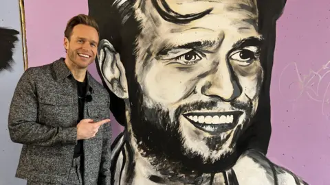 John Fairhall/BBC Olly Murs, a man wearing a grey jacket and trousers and a black top, points at a mural of himself. He has brown hair in a quiff style and is smiling at the camera. The mural is large, black and white, and against a lilac background.