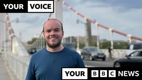 Lewis Eager, young man in blue t-shirt standing in front of a suspension road bridge with cars in the background, with Your Voice Your BBC News branding written around him