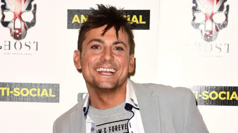 PA Media Paul Danan pictured smiling at a film premiere in London in 2015, he is wearing a grey T-shirt and grey blazer and has short dark hair.