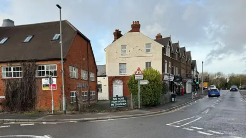 The centre of the village of Tongham. The Kiln building can be seen. There is also another sign saying 'NO MORE NEW HOMES IN TONGHAM.' There is a parade of shops and cars are driving along The Street.