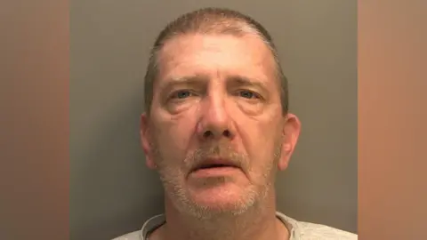 A police mugshot of a man with short brown hair and moustache and grey beard stubble. He is wearing a grey top and looking directly into the camera against a dark wall.