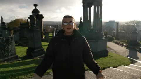 Woman in graveyard with glasses and brown jacket
