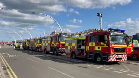 Four fire engines line up along a road outside the amusement park. 