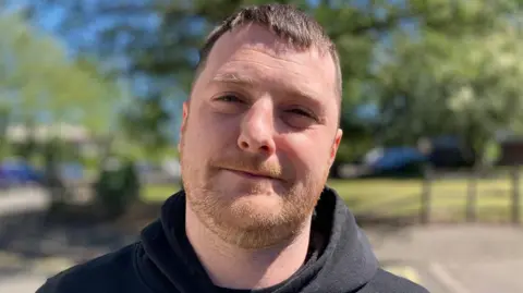 A head and shoulders photograph of Reece Adair. He is wearing a black hooded top.