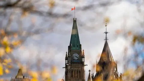 Fall leaves surround the Peace Tower on Parliament Hill. It has a green roof and features a large clock. At the top of its roof is a Canadian flag. 