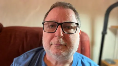 Danny Mendoza sat at home, looking at the camera. He is sat on a red sofa, and is wearing a blue t-shirt and reading glasses. He has short dark hair and a short white beard.