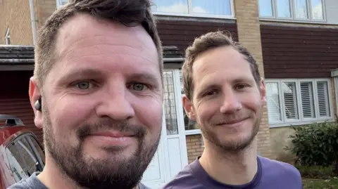 Gareth Hopkins Two men, outside a house, both with short dark hair and beards. They are smiling and looking at the camera. Gareth has an earbud in his right ear. 