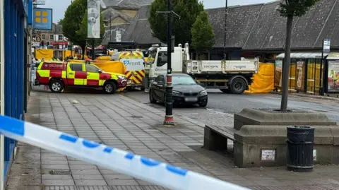 BBC A street which has been cordoned off with police tape and there are several vehicles within the cordon, including trucks and a fire service vehicle.