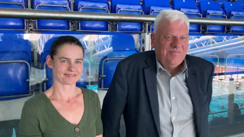 Dr Sarah McNulty has brown hair tied back and is wearing a green top. Graham Morgan has grey hair and and is wearing a grey shirt and blue suit jacket. Both are stood at the side of a swimming pool.