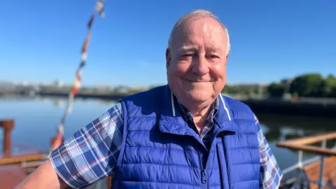 Douglas McGowan, a smiley man with white hair, poses on the Waverley, blue skies and water behind him. He is wearing a blue checked shirt and a royal blue gilet.