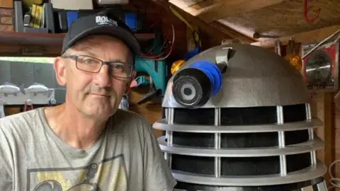 A man wearing glasses and a sun cap and a  grey shirt. He is perched next to a handmade Dalek 