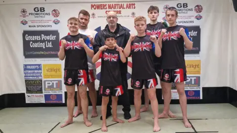 Six martial arts athletes and coach pose with raised fists in a gym, wearing matching ‘Powers-Mac’ uniforms in front of sponsor banners.