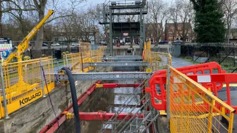 Work being carried out on a lock in Bedford, along a river. A guillotine lock can be seen at one end, with metal items in place, fencing, barriers, and houses behind it all. 