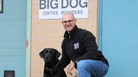 Huw Williams Huw Williams kneels down with his dog, Clyde, who is a black Labrador. The coffee shop signage can be seen in the background, which reads: Big Dog Coffee roasters". Huw wears a black fleece with the logo on the right hand side of his jacket, while clyde sits next to him. A blue door and blue shutters can be seen behind him. 
