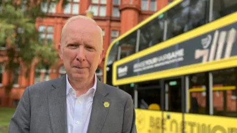 Vernon Everitt, wearing a grey blazer over a white shirt, stands in front of a yellow double-decker bus 