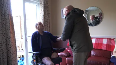 A man in a wheelchair shakes hands with a man stood up. They are in a living room with a red sofa