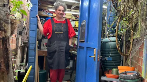 Jackie stands next to a bright blue door, behind her is packed with shelves of her leather, wood and other materials 