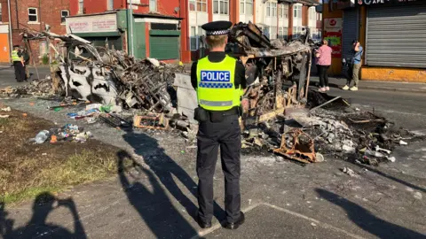 Steve Jones/BBC A police officer guards the scene of a burned out bus