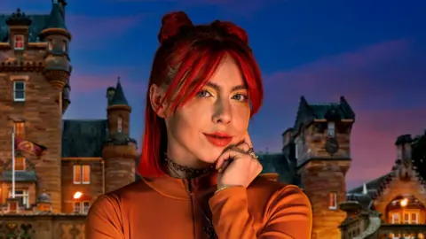 BBC/PA Jessie Stride in her promotional image for The Traitors Series 3. she is wearing an orange top and has red hair. it is a close up image showing her head, shoulders and arm. in the background there is a picture of the traitors castle