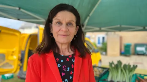 Bridget Smith smiles at the camera. She wears a red jacket and a black top with a red, blue and purple pattern on it. Above her is a green canopy and a number of vegetable boxes.