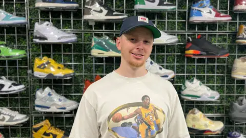 Young boy with cap on stands infront of wall of trainers 