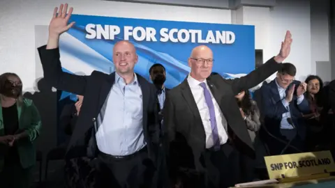 PA Media Stephen Flynn and John Swinney, both bald and wearing dark suits, stand beside each other at an SNP campaign event, each with an arm aloft waving in front of clapping colleagues and a blue SNP billboard. 