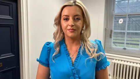 Órlaithí Flynn is standing in a room. A radiator, a window and and door are behind her. Her blond hair is in waves. She's wearing a blue dress. Her silver necklace has a star pendant on it.