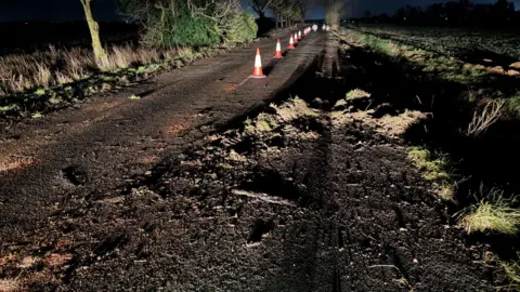 Cambs Police Part of a very muddy and wet road has orange and white cones alongside it