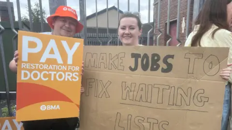 Hannah Patterson holding a large cardboard sign saying 'Make jobs to fix waiting lists'. She has brown hair. She is standing beside another protester who has an orange BMA had and is holding an orange sign saying 'Pay restoration for doctors'.