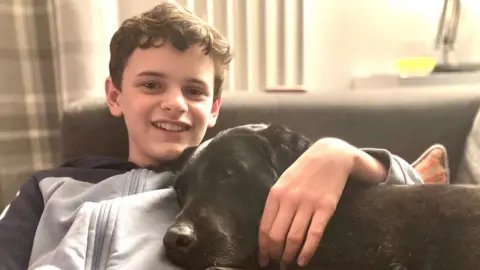 A boy in a light-coloured hoody with a dark stripe on the arms and shoulders is sitting on a sofa and hugging a dog. The boy is smiling and looking at the camera