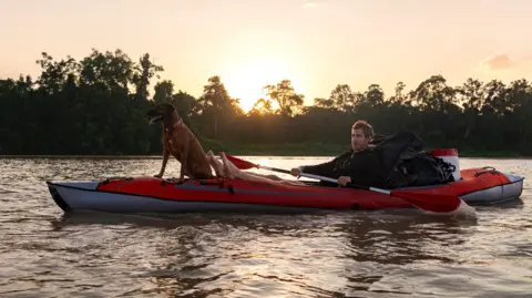 Jacob Hudson Ash Dykes in his kayak with his feet up and with the dog sitting at the front as the sun sets