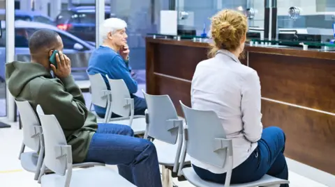 Patients waiting in chairs in a hospital A&E unit: they are seated in rows - two sit in the front row, a man on his phone is seated in the row behind them (library image). 