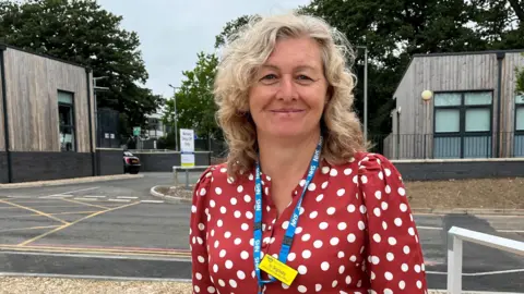 Clare Worden/BBC Jo Segasby wears a red-and-white spotted blouse and stands next to hospital buildings.