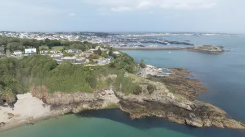 BBC Fort George from the sky - A cliff face from the sky, with leafy green growth on the side of the cliff face and some houses atop it. 