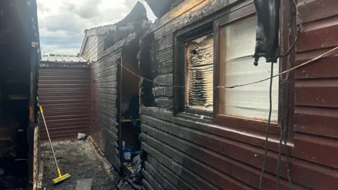 The side of a wooden building affected by fire. A window and blinds inside it are charred and broken, and a door and part of its roof has been burnt and damaged