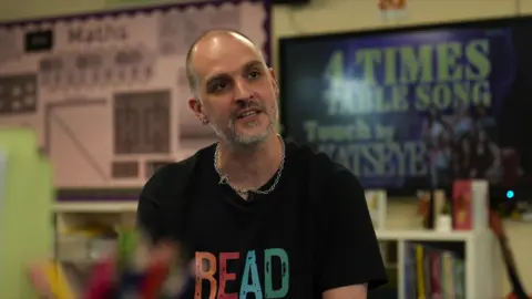 Chris Walker is bald with a short greying beard. He has a small hoop earring, a neck chain and a black T-shirt with the word "READ" written across it in bold capitals.