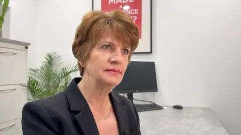 A woman with short brown hair is wearing a black blazer. She is sat at a desk with a computer monitor. There is a plant and a filing cabinet behind. 
