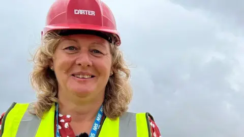 Woman with hard hat and high-vis vest stands infront of a metal fence. The fence is sectioning an area which has been demolished. A building is in the background.
