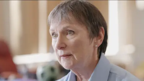 Cwmni/S4C Lee McKenzie is a woman in her 60s with short greying dark hair. She is wearing a light blue shirt with a collar. Her head and shoulders can be seen. She is looking off to the left of the picture.