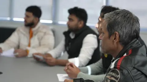 Four men sit at a round table in an office looking sideways towards a BBC reporter