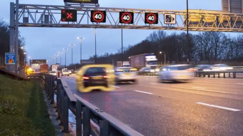 West Mercia Police Cars are driving at speed down a motorway, under a signage gantry. Above the car in the far left lane there is a red "X" sign, which means that the lane cannot be used.