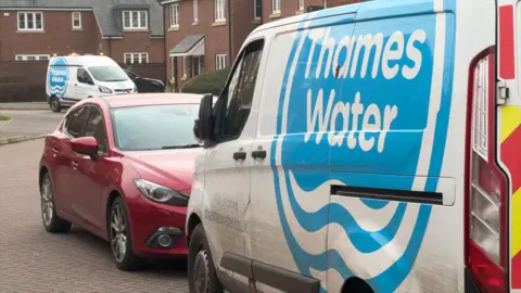Two white vans, with the bright blue Thames Water logo on the sides, are parked-up on a residential street.