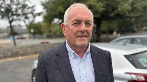 Fianna Fáil TD John McGuinness, with short white hair, wearing a blue shirt and dark grey suit. There is a silver car parked behind him. 